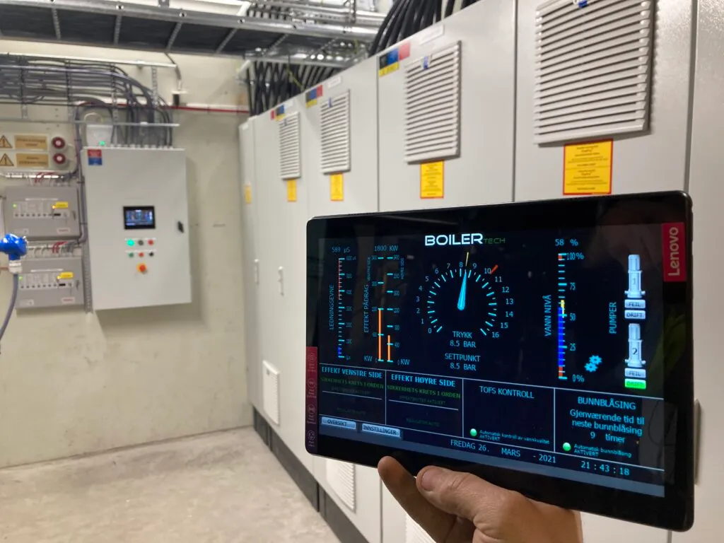 A boiler room with control cabinets; a hand holds a tablet showing boiler pressure, output and water level